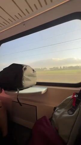 The video shows the interior of a train during the day. In the foreground, a black backpack rests on a small shelf by the window. Outside the window, a sunny landscape with fields and utility poles is visible. Inside the train, two young women are seated next to each other. The woman on the right wears a red shirt and is looking down at her phone. The woman on the left wears a purple and white patterned top and white shorts, and she is also looking at her phone. In the background, other passengers are seated on the train, some looking towards the front of the train. The train carriage has blue seats and yellow overhead lighting.