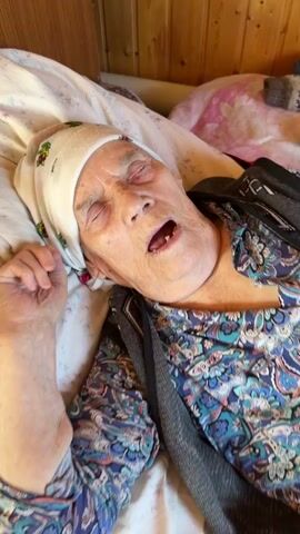 The video shows an elderly woman lying in bed with her eyes closed and mouth open. She is wearing a headscarf and a patterned top. Her left arm is visible, resting on the bed. The background appears to be a room with wooden walls. There are no other people or significant objects visible in the frame. The woman's facial expression suggests she might be sleeping or unwell.