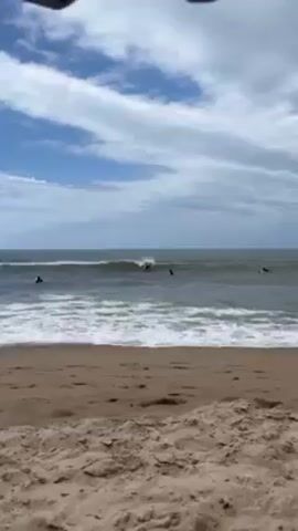 The video shows a beach scene with several people surfing in the ocean. The beach has light brown sand, and the ocean waves are choppy with white foam. The sky is mostly cloudy. One surfer, dressed in black, rides a red surfboard. He is shown standing with his arms outstretched, before falling off the board. Another surfer is seen sitting in the water nearby. The camera view is static, positioned on the beach.