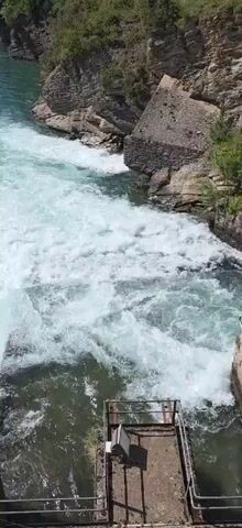 The video showcases a river with turbulent, white water flowing between rocky banks and green, tree-covered hills. The river's color transitions from a deep blue-green to a foamy white, indicative of rapid movement and aeration. Concrete structures line parts of the riverbank, suggesting human intervention or modification to the natural landscape. A small metal apparatus, possibly part of a weir or similar water control mechanism, is visible at the bottom of the frame.