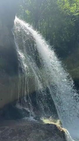 The video shows a waterfall cascading into a pool of water. The waterfall is surrounded by lush green trees and rocky cliffs. The water in the pool is a light green color. The sun is shining brightly, creating a sparkling effect on the water. The camera pans from the top of the waterfall to the pool below.