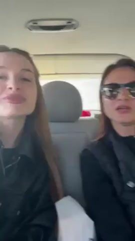 Two women are sitting in a car. The woman on the left has long brown hair and is wearing a black jacket. She is smiling and looking at the camera. The woman on the right has long brown hair and is wearing sunglasses and a black vest. She is also looking at the camera. The woman on the left holds her hand up to show off her bright red fingernails. Music is playing in the background.