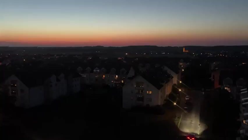 This aerial video captures a dusk scene over Mühldorf a. Inn, Germany. The sky displays a gradient of colors from a deep blue at the top to a vibrant orange and pink band near the horizon, indicating the transition from day to night. Below, a suburban or town area is visible, characterized by rows of multi-story residential buildings. Many of these buildings have lit windows, suggesting residents are home as evening sets in.

A street winds through the buildings, illuminated by streetlights. A red car with its headlights on is driving down this street, its taillights visible as it moves away from the camera. The overall impression is one of a quiet, peaceful evening in a residential community. The terrain appears somewhat hilly, with the distant landscape fading into a hazy silhouette against the colorful sky. No people are clearly discernible, and no text is visible in the footage. The weather appears clear, with no signs of rain or heavy clouds. The time of day is clearly dusk, as evidenced by the dramatic sky and the artificial lighting appearing in the buildings and on the street.