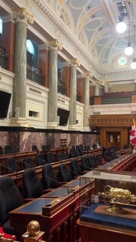 The video shows the interior of a legislative chamber. Rows of dark upholstered seats with wooden desks and built-in microphones are arranged in a horseshoe shape facing a raised central dais. The chamber features ornate classical architecture with tall marble columns, gilded detailing, and a highly decorated vaulted ceiling with circular windows and light fixtures. Two flags are visible on the dais: a Canadian flag and a provincial flag. In the foreground, a golden ceremonial mace is displayed under glass. The room appears empty of people.