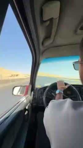 A man drives a red car on a highway. The car is older, and the interior looks worn. The road is flanked by hills, and the sky is clear. The man is wearing sunglasses and a watch.