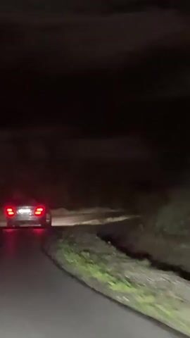The scene is a nighttime view of a car driving on a dark, winding road. The car's rear taillights are illuminated, and it appears to be moving away from the viewer. The road surface is wet, reflecting the taillight's glow. The sides of the road are lined with vegetation. The sky is dark and partially obscured by clouds.
