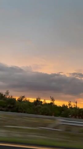The video shows a blurred view of a sunset from a moving vehicle. The sky is a mix of soft orange and gray clouds. Below the sky, there is a line of trees and vegetation. A highway barrier is visible in the foreground, indicating the perspective is from a car traveling on a road. The overall impression is one of a scenic drive during dusk.