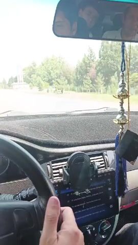 A first-person perspective video shows the interior of a car. The camera is mounted on the dashboard facing forward. The steering wheel and dashboard are visible in the foreground. A hand is on the steering wheel. A phone is mounted on the dashboard, displaying a music player interface. In the rear-view mirror, two people are visible. Outside the car, trees and a monument or sign are seen. The car appears to be moving on a road.