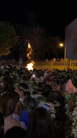This video captures a lively nighttime outdoor gathering. A large crowd of people, many wearing floral wreaths around their necks or heads, are packed together. In the background, a bonfire burns brightly, casting an orange glow and sending sparks upwards. Palm trees are visible against the dark night sky, and a street lamp illuminates part of the scene. The crowd appears engaged, with some holding up phones, likely recording or taking pictures of the bonfire and festivities. A decorative pink fan is also visible amongst the people. The overall atmosphere suggests a celebratory event or festival.