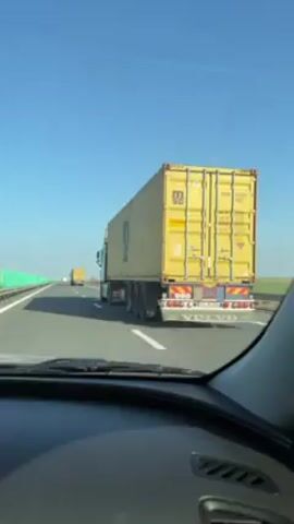 The video shows a car driving on a highway with two lanes in each direction. A large truck carrying a yellow shipping container is in the right lane.  The truck has the letters "MSC" written on the container. Another yellow truck is visible far ahead in the same lane. A green noise barrier lines the left side of the highway. The sky is clear and blue. The dashboard of the car is visible at the bottom of the frame. The license plate on the first truck is visible and reads "B 200 BA".