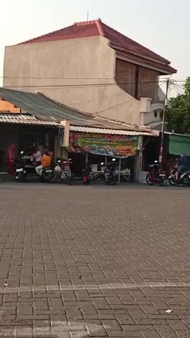 The video shows a street scene in what appears to be a residential or commercial area. The ground is paved with interlocking bricks. On the right side of the street, there are several shops with awnings and motorcycles parked outside. One shop has a banner with text in Indonesian. The buildings are mostly single or two-story structures with tiled roofs. Indonesian flags, red and white, are prominently displayed on poles, some of which are quite tall and appear to be temporary installations, possibly for a celebration or event. In the background, more buildings and trees are visible, and the sky is bright, suggesting daytime. Several motorcycles are seen moving on the street, and a white minivan is parked on the left. The overall impression is a bustling street in a Southeast Asian country, likely Indonesia, given the flags.