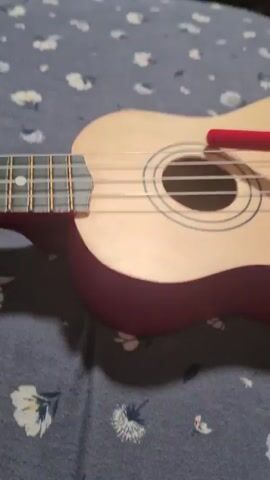 A close-up shot focuses on a ukulele, its strings and fretboard visible. A person's hand is seen holding a red object, possibly a pick or a stylus, near the soundhole. The ukulele rests on a surface covered with a blue fabric featuring a floral pattern. The camera pans slightly, capturing a portion of the ukulele's body. The overall scene is dimly lit, with the red object creating a sharp contrast against the instrument.