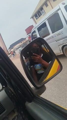 A person is taking a selfie in the side mirror of a vehicle. The person is holding a phone and looking at it. The vehicle appears to be in traffic. In the background, there are other vehicles and buildings. The sky is overcast.