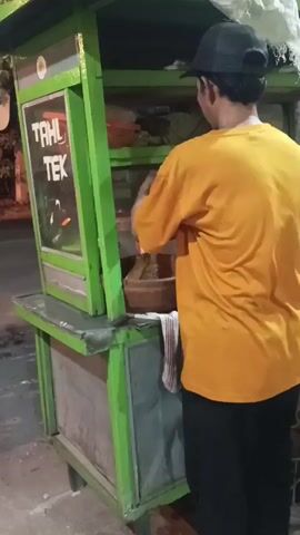 A person wearing an orange t-shirt and a black baseball cap stands behind a green food cart with "TAHU TEK" written on it. The cart has shelves filled with various items. The person is engaged in preparing food, possibly using a mortar and pestle. The cart has wheels and appears to be on a street or sidewalk. The lighting suggests it is evening or nighttime.