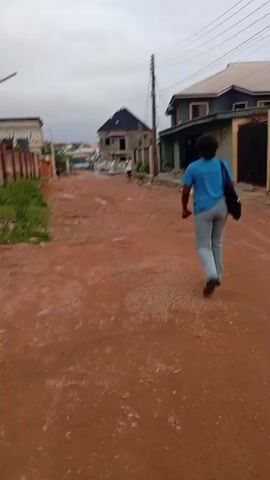 This media file captures a street scene in Idimu, Nigeria, with a cloudy sky suggesting an overcast day. The street itself is unpaved and appears to be made of red dirt, with some areas showing signs of being muddy or waterlogged. The ground is uneven and strewn with gravel and debris.

The primary subject is a person walking away from the camera, dressed in a blue t-shirt and grey trousers, carrying a dark bag over their shoulder. Their gait suggests a casual pace. In the background, there are several houses under construction or recently built. One prominent house in the middle ground is a multi-story structure with a dark, angular roof. To the right, another house is visible, painted in muted tones of green and grey, with a black gate. Further down the street, other buildings and the general landscape of a developing urban area are visible.

A solitary person is also visible in the distance, walking along the street. A utility pole with wires stands tall on the right side of the frame, indicating the presence of infrastructure. The overall atmosphere is somewhat subdued, likely due to the cloudy weather and the unfinished state of some of the buildings, which hints at ongoing development or a less-than-pristine environment. There is no visible text in the media file. The time of day is unclear but appears to be daylight.