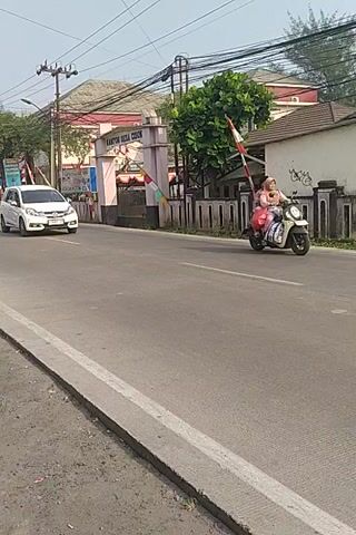 The video shows a white car driving on a paved road. A motorbike is following closely behind the car. The road is lined with a concrete curb and a grassy area. There are buildings and trees on the left side of the road. The sky is visible at the top of the frame. The video is shaky, suggesting it was recorded from a moving vehicle.