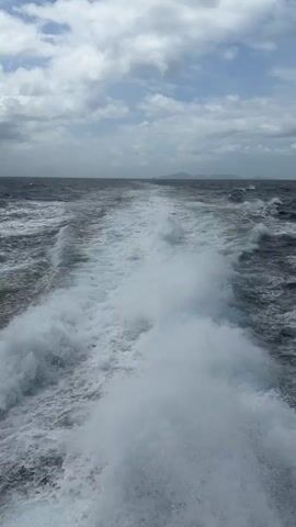 This is a first-person perspective video of a boat moving through the ocean, creating a large wake. The water is choppy with white foam. In the distance, there is a hazy horizon with what appears to be land or islands. The sky is partly cloudy with patches of blue. The camera is pointed backwards, showing the disturbed water trailing the boat. There are no people visible in the video.