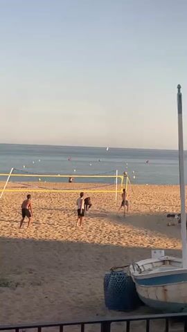Four shirtless men are playing beach volleyball on a sunny day. The volleyball net is strung up on a sandy beach. The ocean is in the background, with several buoys visible in the water. A partially visible boat rests near the bottom right of the frame, next to a large, woven, blue basket. The video is shot from an elevated, possibly indoor, position, with a railing partially visible at the bottom of the frame.