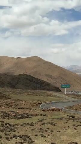 The video presents a scenic view of a mountainous landscape, likely in a high-altitude region. The mountains are predominantly brown and tan in color, with rocky terrain visible in the foreground. The sky is partly cloudy, with patches of blue visible between the white clouds. A road winds through the landscape, with a sign visible on the roadside. The overall environment appears arid and sparsely vegetated.
