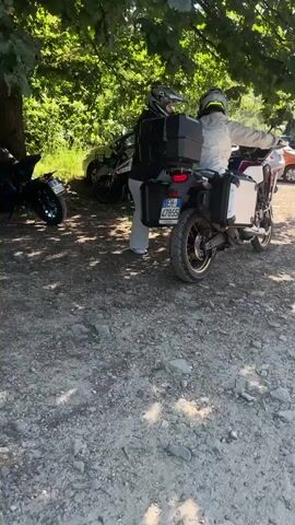 The video shows a gravel parking lot on a sunny day. Two people in motorcycle helmets are preparing a touring motorcycle with storage cases. The motorcycle is mostly white with red and gold accents. Other parked motorcycles and cars are visible. The camera pans to show a group of four women walking away from the motorcycle. One woman is wearing white pants and a white top and carrying a large white bag. Buildings and trees are in the background.