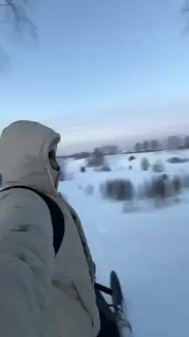 The video shows a person riding a snowmobile through a snowy landscape. The person is wearing a light-colored hooded jacket and a backpack. The snowmobile is white with red accents. The landscape is flat and covered in snow, with sparse trees. The sky is a clear blue with a pink and orange glow on the horizon. A crescent moon is visible in the sky. The camera is mounted on the snowmobile and is moving forward. The video is slightly blurry and shaky.