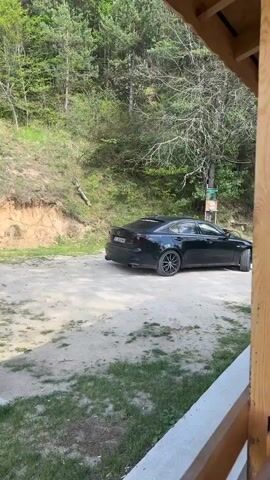 From an interior vantage point, the video opens with a view of a black car parked in a gravel lot, framed by the wooden beams of a structure. The camera pans to the left, revealing a large, wooden heart-shaped structure, followed by a vista of lush green trees. The camera then tilts upwards, showcasing a breathtaking mountain range partially covered with snow, set against a backdrop of a blue sky dotted with fluffy white clouds.