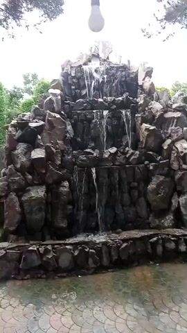 The video shows a multi-tiered rock waterfall. Water cascades down the rocks into a pool at the bottom. The waterfall is surrounded by greenery and trees. A lightbulb hangs from a wire above the waterfall. The ground in front of the waterfall is paved with a circular pattern. The video is shot from a medium distance.