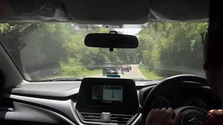 The video is filmed from the perspective of someone driving a car. In the foreground, the steering wheel and dashboard are visible. The car's touchscreen infotainment system displays a navigation map. The windshield shows a two-lane road winding through a lush, green forest. Several vehicles are visible on the road ahead, including a black SUV, a red car, and a motorcycle. The sun appears to be shining, casting dappled light through the trees. A person's hands are on the steering wheel, suggesting they are driving the car.