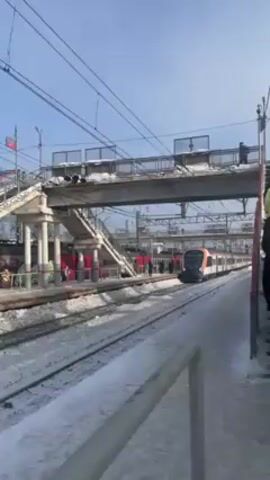 The video shows a modern commuter train arriving at a snow-covered platform under a pedestrian overpass. The train is white with red and orange stripes, and it approaches from the right side of the frame. The platform and tracks are visibly covered in snow, and the sky is a clear blue.  A few people are visible on the overpass. The camera is stationary, focusing on the train as it comes to a stop.