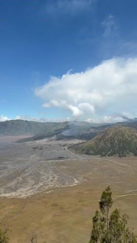 The video opens with a wide shot of a vast, arid valley with a light brown, sandy terrain. In the distance, a volcanic crater emits a plume of smoke, with greener, mountainous terrain surrounding it. The sky is a clear, vibrant blue with scattered white clouds. As the video progresses, the camera zooms in on the volcanic crater. The crater's rim is jagged and appears to be composed of dark, rocky material. The sides of the crater are steep and textured, with layers of ash and rock visible. The smoke billowing from the crater is thick and grey, obscuring some of the interior. In the foreground of the zoomed-in view, there is a flat, dark area, possibly a parking lot or a base camp, with a few small buildings and some vehicles. The surrounding mountains are covered in green vegetation, with shadows cast by the sun, indicating the time of day. The terrain in the valley appears to be a dried-up riverbed or a volcanic ash plain, with intricate patterns formed by the flow of water or ash. The overall impression is a majestic and somewhat desolate volcanic landscape under a bright, sunny sky.