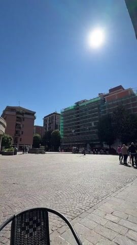 The video shows a sunny day in an Italian city square. The camera pans from a near view of the empty brick-paved square to an outdoor cafe with red awnings and several patrons seated at tables. A few people are walking across the square. A large building is visible across the square covered in scaffolding and green mesh. The sun is bright in a cloudless sky.