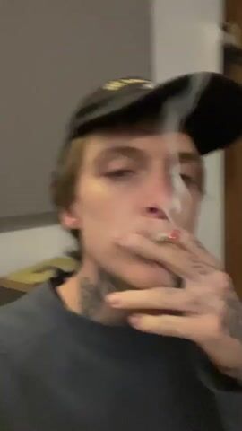 The video shows a young man, viewed from the chest up, smoking what appears to be a marijuana cigarette or joint. The man is wearing a dark-colored baseball cap and a dark-colored crew-neck sweater. He has fair skin and neck tattoos. The focus is on the man's face as he inhales and exhales smoke. The background is blurred, but a light-colored wall and a framed picture can be seen. The man brings the joint very close to the camera at the end of the video.