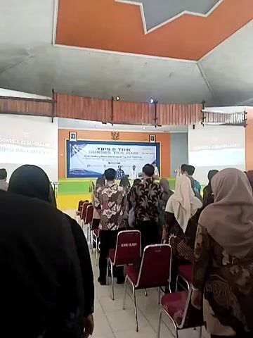 The video shows an indoor event with a stage at the far end of a room. A banner on the stage reads "TIPS & TRIK SUKSES TKA 2026 PEMBUATAN AKUN DASHBOARD TRY OUT YUINTER". Several people are standing on the stage, and a few are seated behind a table. In the foreground and middle ground, a group of people are seated in red chairs, facing the stage. Most of the people visible are wearing traditional Indonesian batik shirts. The room appears to be a conference hall or auditorium, with wooden paneling on the ceiling and orange accents on the walls. The lighting suggests it is daytime, but the interior is illuminated by artificial lights. The camera is positioned behind some of the seated attendees, giving a view over their heads towards the stage. The overall atmosphere appears to be formal, possibly a seminar or workshop.