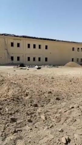 The video shows a building with damage to the roof and walls. There are numerous holes in the building, and some debris is visible. In front of the building, there is a large crater in the ground, suggesting an explosion. The surrounding area is barren and desert-like. In the distance, there are other buildings that appear to be similar in style. People are walking around the crater. The sky is clear and blue, and the sun is shining brightly.