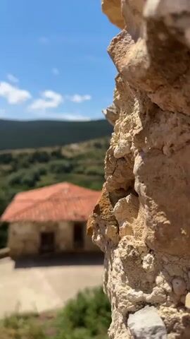 The video shows a view of a rural landscape through a stone wall. In the foreground, a rough stone texture dominates the right side of the frame. Out of focus in the middle ground, a small stone house with a red-tiled roof is visible. Behind the house, rolling hills covered in greenery and trees stretch towards the horizon. The sky is blue with a few scattered white clouds. The overall scene depicts a sunny day in a picturesque, possibly ancient, village setting.