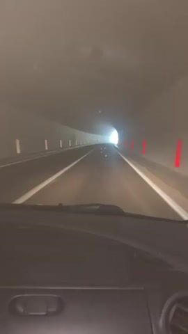 The video shows a first-person view from inside a car driving through a tunnel. The tunnel is dimly lit, with a bright light visible at the end. The road ahead is dark asphalt with white lines marking the lanes. On either side of the road, there are vertical white markers and orange-red bollards. The walls of the tunnel appear to be concrete and are wet, possibly due to condensation or water. Small specks, possibly dust or water droplets, are visible in the air, reflecting the light. The dashboard of the car is visible in the foreground. The car appears to be moving at a moderate speed.