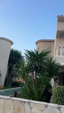 The video pans across the exterior of a residential building. The building appears to be a multi-story house or apartment complex with cream-colored walls and a tiled roof. It features balconies with white railings. Lush green plants and some flowering bushes with pink blossoms are visible in front of the building and along the side. A stone wall runs along the foreground. The sky is clear and blue, suggesting it is daytime. Further down the street, cars are parked along the sidewalk, and other similar buildings can be seen. There are no people visible in the video.