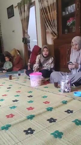 A video shows a group of people sitting on a woven mat inside a room. The room has brick-patterned walls and curtains. A television is visible on the left. In the foreground, someone's hand is holding a phone, recording the scene. In the background, a woman in a red hijab sits on the mat, facing the camera. To her left, another woman in a brown hijab is seated. A young child in a light green shirt is also on the mat. Further back, a woman in a dark hijab walks through a doorway. A pink container with snacks sits on the mat. There are also cups of tea visible.