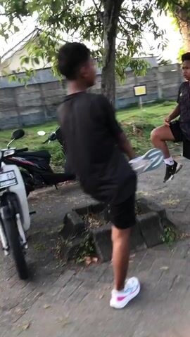 The video shows two young men in an outdoor setting. The foreground features one man wearing a black t-shirt and black shorts, engaged in what appears to be a stretching exercise with a white and blue object. He is also wearing white sneakers with pink and blue accents. A white motorcycle is visible to his left. In the background, another young man is seated on a low wall, wearing a dark t-shirt and black shorts with white socks and dark sneakers. The environment seems to be a paved area with a tree and a fence in the background. The lighting suggests it is daytime. The video is slightly blurry, indicating motion.