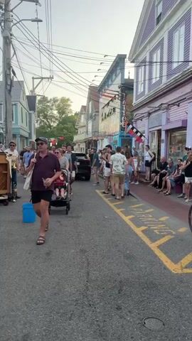 A street performance is taking place on a paved street lined with buildings. A band is playing instruments, including a piano, drums, and a brass instrument, on a makeshift stage on the left side of the street. A crowd of people watches the performance and walks down the street. Some people are pushing strollers, and others are holding drinks. There are power lines and traffic lights visible overhead. The buildings appear to be shops or restaurants, with some decorated with flags and banners. The sky is overcast. The overall atmosphere seems festive and lively.