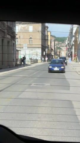 The video shows a street scene in Sarajevo, Bosnia and Herzegovina. The camera is positioned inside a vehicle, looking out through the rear window, which has defroster lines visible. The time of day appears to be daytime, with bright sunlight casting shadows on the street. The weather seems to be clear and sunny.

The scene depicts a street lined with old buildings, some of which appear to be commercial establishments. One building has a red sign with white text that reads "OPTIKA". There are also signs that appear to be for "FRANC" and "OPTIC".

Several people are visible walking on the sidewalk. Two cars are driving on the street: a blue Volkswagen Passat station wagon is in the foreground, and a dark-colored SUV or van is further behind. The street has visible road markings, including yellow lines and what appear to be tram tracks. In the background, a hillside with buildings is visible, suggesting the city is situated in a valley. The overall impression is of a typical urban street scene on a pleasant day.