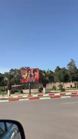 The video shows a street view with a park on the right. The day is bright and sunny with a clear blue sky. There are palm trees and other trees in the park. People can be seen walking in the park. There are billboards along the street. The street has a red and white curb. There are cars passing on the road.