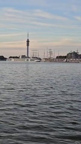 A wide shot shows a body of water in the foreground with gentle waves. In the background, a city skyline is visible under a cloudy sky. A tall, slender tower with a pointed top stands out prominently on the left side of the skyline. To the right of the tower, several large ships, including at least two multi-masted sailing vessels, are docked. A long, modern white vessel is positioned between the tower and the sailing ships. Buildings with windows and balconies line the waterfront behind the docked ships. The overall scene suggests a harbor or port city. The video appears to be static, with no discernible movement other than the natural ripple of the water.