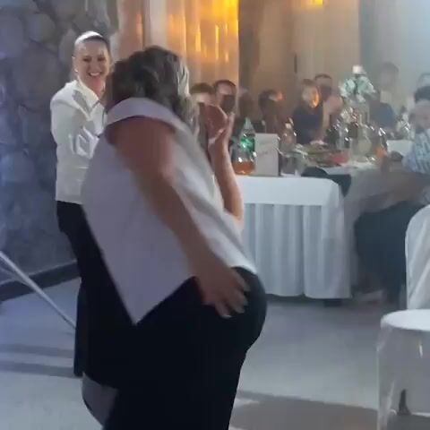 The video shows a group of people at what appears to be a wedding reception. In the foreground, a woman is being hugged from behind by a man who is on one knee. The woman has her hands on the man's shoulders. Behind them, a woman in a white blazer and a ponytail is singing into a microphone. In the background, there are tables set with white tablecloths, food, and drinks. Other guests are seated at the tables, watching the events unfold. The environment is dimly lit with warm spotlights.