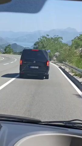 The media shows a first-person perspective from inside a vehicle, looking forward. The camera is positioned behind the dashboard, with a clear view of the windshield and a portion of the interior. Ahead, on a winding, paved road, a black Mercedes-Benz Vito van is traveling in the same direction. The van has its taillights illuminated. The license plate of the van is visible and reads "BD BG734".

To the left of the road, there is a steep, vegetated slope with trees and rocks. In the distance, a body of water, likely a lake or sea, is visible, bordered by rolling hills and mountains. The sky is clear and blue, suggesting a sunny day. A blue road sign with a white "P" symbol is present on the right side of the road.

The overall scene depicts a scenic drive on a sunny day, possibly in a mountainous or coastal region of Montenegro, given the location context provided. The activity is simply traveling by car on a road with other vehicles. The lighting indicates it is daytime. There are no discernible emotions, but the clear weather and scenic view suggest a pleasant journey.