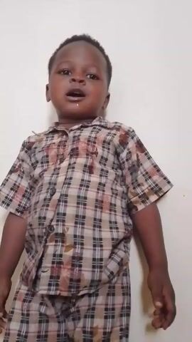 A toddler stands against a white wall. He wears a brown and black plaid button-up shirt and matching pants. He appears to be speaking, with his mouth open. He then claps his hands together.