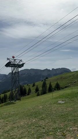 The video shows a scenic mountain landscape under a partly cloudy sky. A tall, metal ski lift tower stands on a green, grassy hillside. Two women wearing backpacks and casual clothes walk along a path towards the tower and then continue, their backs to the camera. The background features rolling green hills and distant mountains. The camera pans slightly to reveal a wider view of the mountainous landscape.