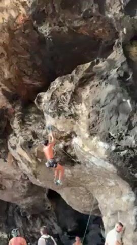 A man is rock climbing up the underside of a light-colored rock formation. He is shirtless, wearing dark shorts and climbing shoes. He is attached to a rope, and there are other people watching from below. The rock formation is varied in texture and color, with some dark sections.