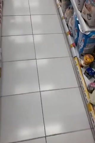The video shows a person walking down a grocery store aisle. The aisle is lined with shelves stocked with various products, including what appear to be diapers and baby formula. The floor is tiled, and the lighting is bright. The person is wearing dark shoes and jeans. The camera is pointed downwards, showing the floor and the shelves. The person is walking from right to left in the frame. There are no other people visible in the immediate vicinity.