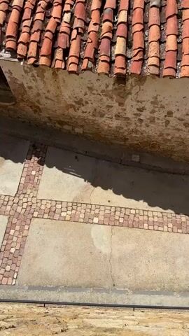 The video begins with a close-up view of a weathered terracotta tiled roof. The tiles are old and some are broken, with moss or dirt accumulated in the crevices. Below the roof, a roughly plastered wall shows signs of peeling paint and damage. The ground in front is paved with small square tiles arranged in a geometric pattern, with shadows cast across it, indicating sunlight.

The camera then pans up and to the right, revealing a wider view of the surrounding environment. In the mid-ground, there are buildings with similar red tiled roofs. The landscape beyond consists of rolling hills covered in dry grasses and scattered trees. In the distance, a dark green, dense forest covers the larger hills. The sky is a clear blue with a few scattered white clouds.

The video continues to pan and zoom out, showcasing more of the village and its setting. More buildings with red roofs are visible, some appearing in better condition than others. A narrow, paved road winds through the village, with a car parked on it. The natural landscape remains prominent, with the hills and forest stretching towards the horizon. The overall impression is of a rural, somewhat aged village situated in a hilly, natural environment under a bright, sunny sky.